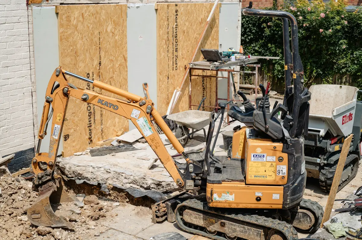Mini-pelle au travail contre une maison, au milieu des déblais et des panneaux de chantier