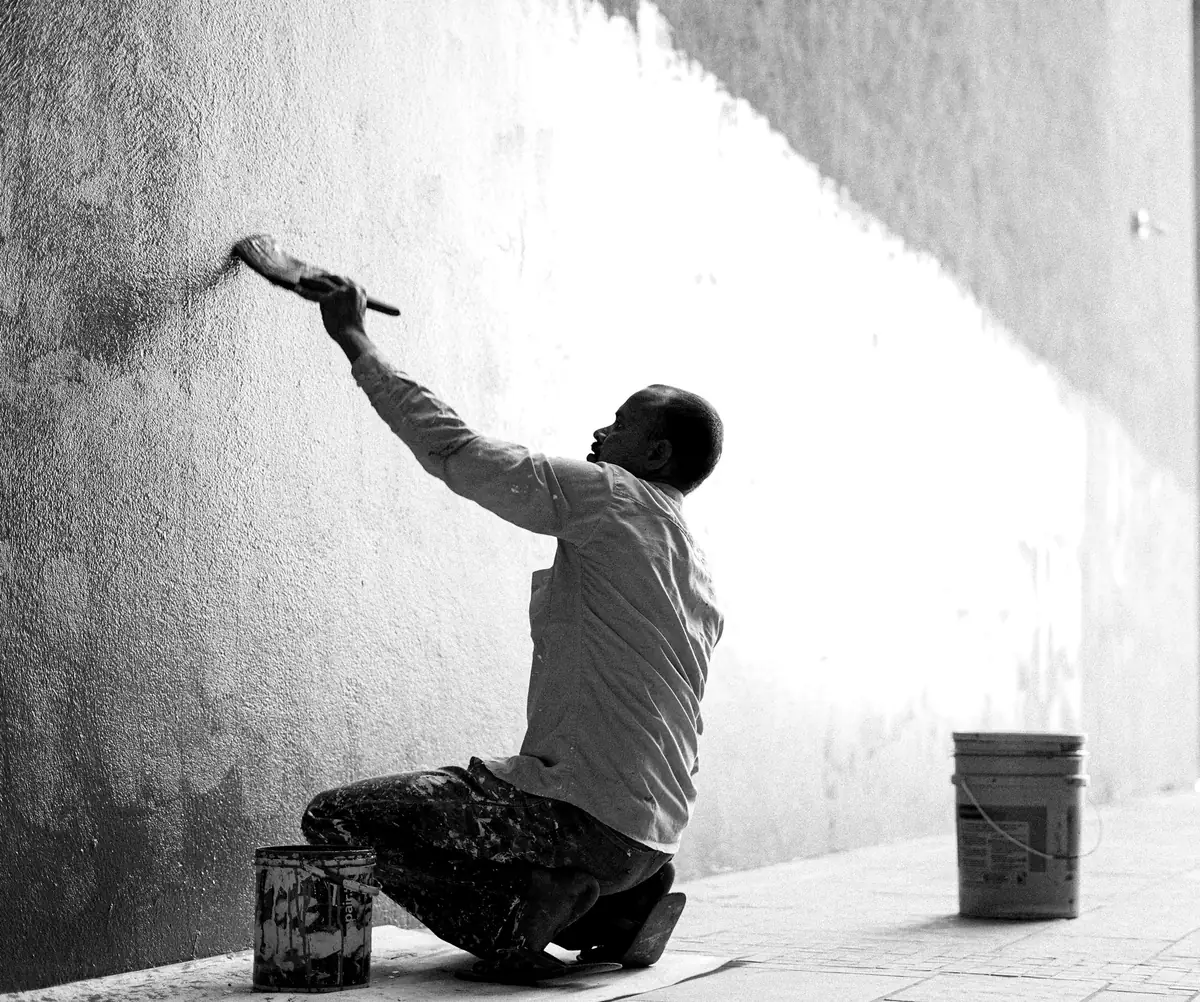 Peintre accroupi qui passe la brosse le long d’un mur blanc, pot à ses pieds