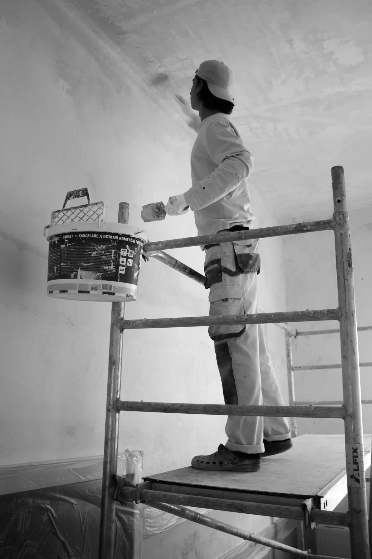 Peintre debout sur un échafaudage roulant, seau à la main, devant un mur clair