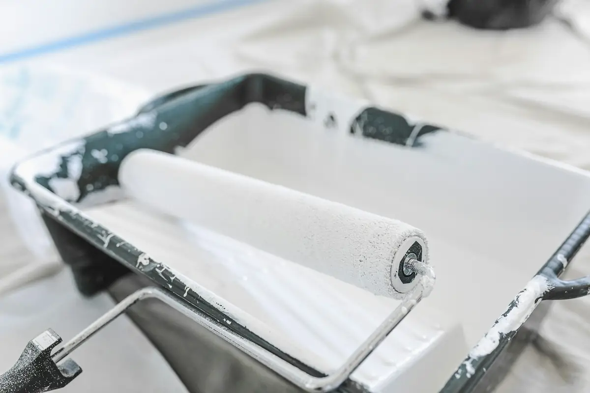 Rouleau de peinture blanc posé sur son bac, bâche de protection au sol
